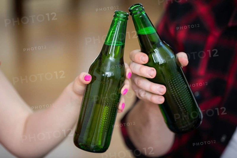 A close-up on the hands of two people. Each of them is holding a green bottle without a label. The boy and girl are giving a toast. Blurred background