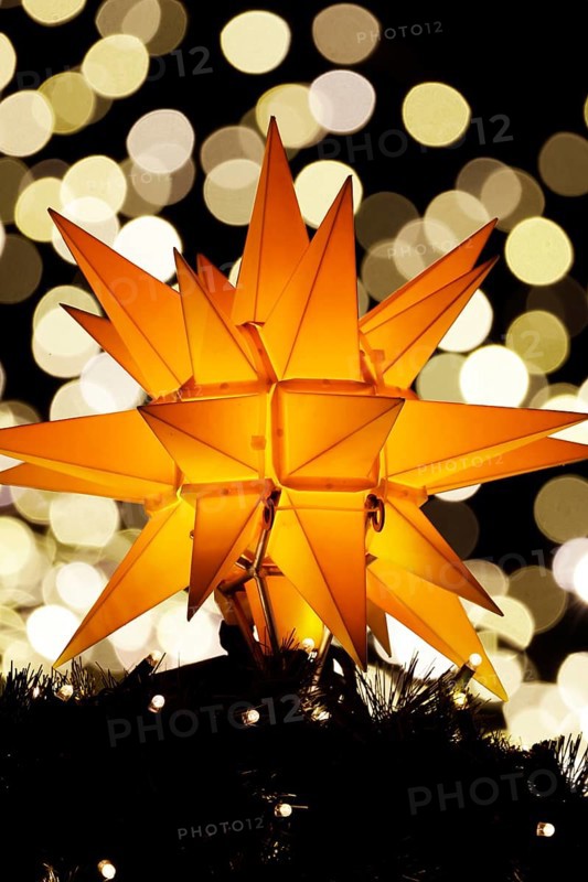 An orange-coloured, Christmassy star shines in the foreground in front of a beautiful, blurred bokeh background with lots of glittering, sparkling lights at a Christmas market at night, Cologne, North Rhine-Westphalia, Germany, Europe