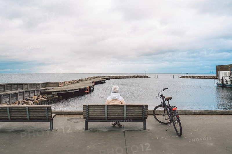 A young Caucasian woman sits with her back on a wooden bench overlooking the Baltic Sea on the seafront in Copenhagen Denmark in winter in cloudy weather. Girl walking gonoskoy bike parked nearby