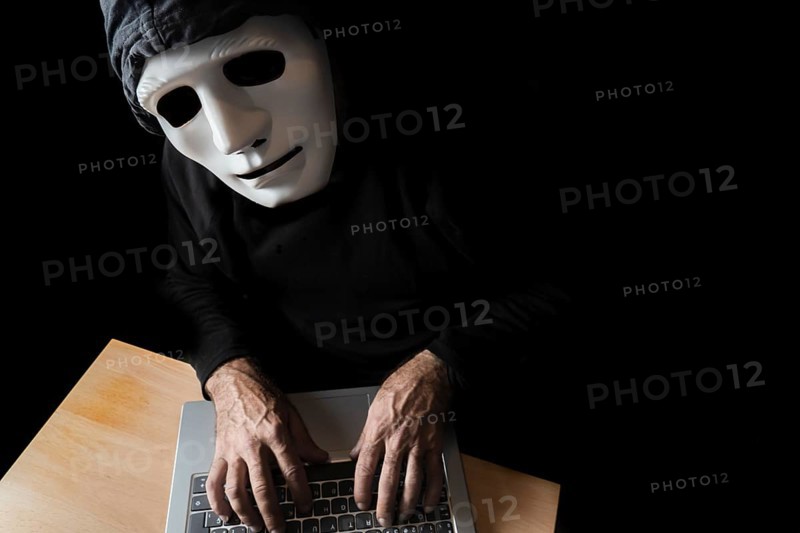 Hacker dressed in black with mask and hood typing on a computer while looking at the camera cybercrime concept