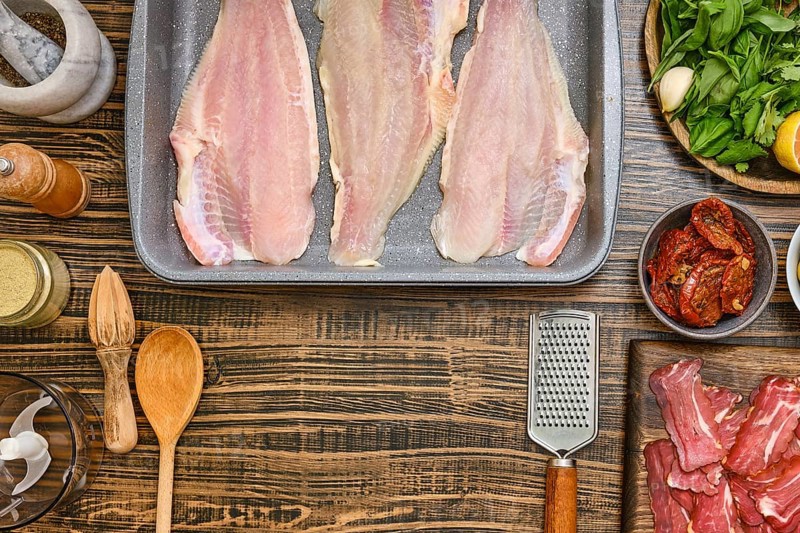 Fresh fish fillets are laid out on a baking tray, surrounded by various ingredients including herbs, lemon, and spices. Wooden utensils and bowls add a rustic touch to the kitchen workspace