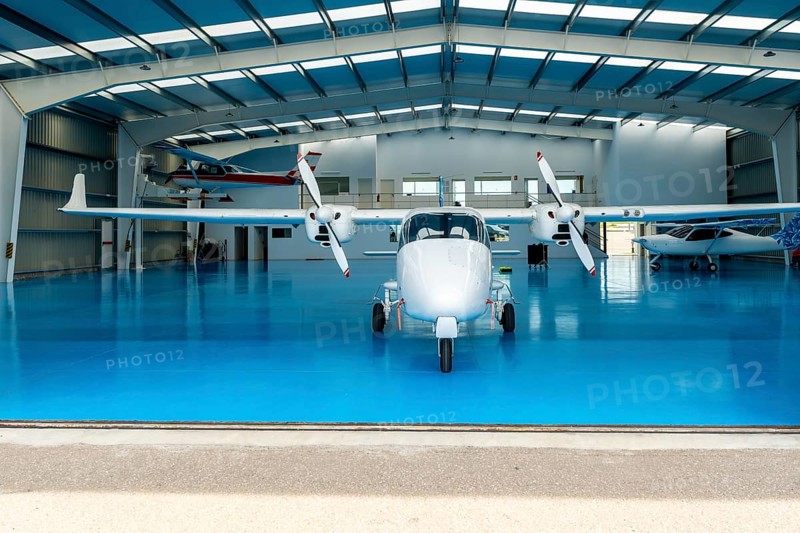 White twin engine airplane parked inside a spacious hangar, featuring a bright blue floor and surrounded by smaller aircraft