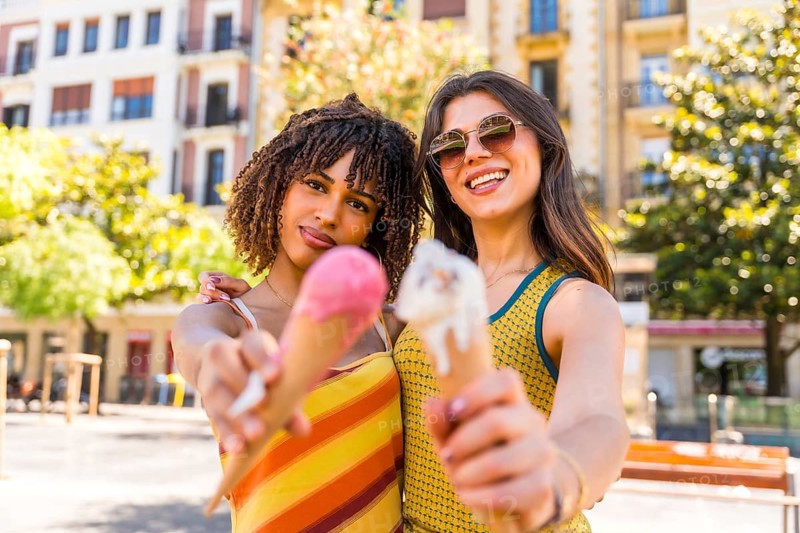 Two friends enjoying a sunny day in the city, happily holding and sharing colorful ice cream cones while smiling in the warm sunshine