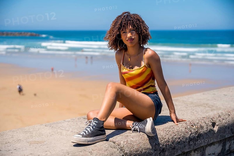 Stylish young woman wearing casual summer clothes sits on a beach wall, enjoying the ocean view during her vacation