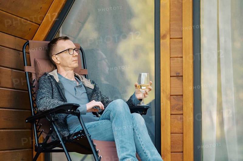 Middle-aged man relax on terrace of a tiny house with a glass of wine