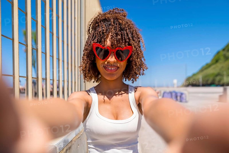 Smiling young woman with curly hair taking a selfie with heart shaped sunglasses on a sunny beach boardwalk