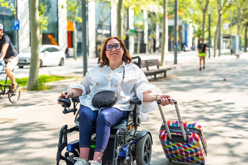 Happy woman with disability enjoying a sunny day outdoors while traveling with her electric wheelchair and colorful suitcase