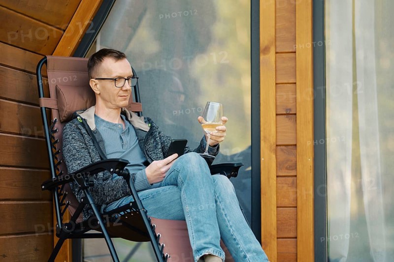 Middle-aged man looking at smartphone sitting on terrace of a house with a glass of wine