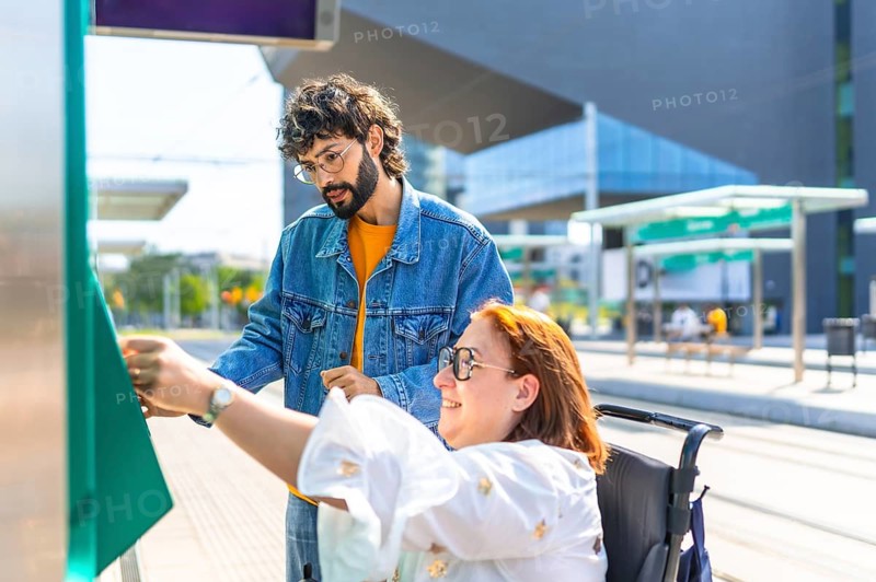 Woman in a wheelchair validating her ticket at a modern tram stop, with a friend providing assistance and support during the process