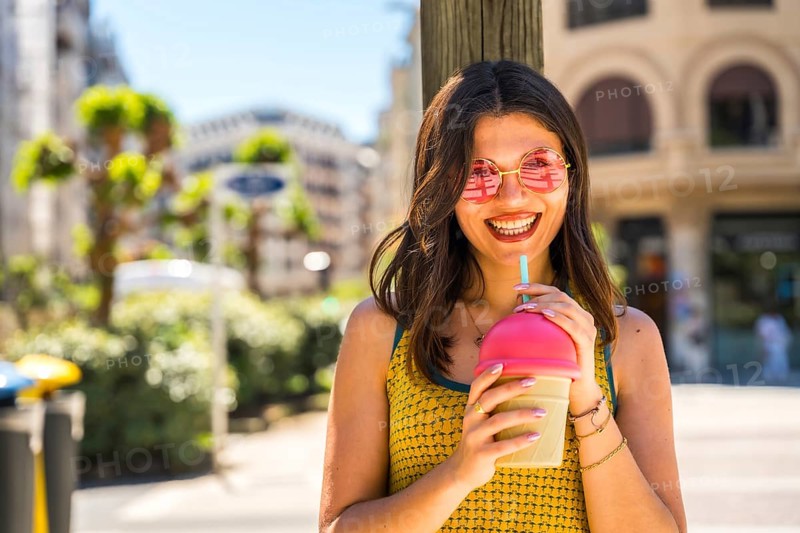 Smiling woman with pink sunglasses enjoying a refreshing fruit smoothie on a sunny summer day in the city