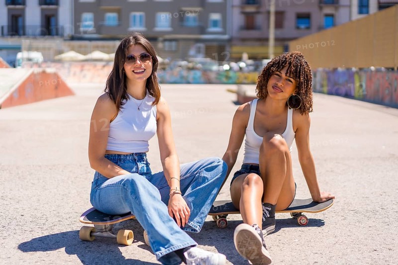 Two young women are sitting on their skateboards in a skate park, enjoying a break and smiling