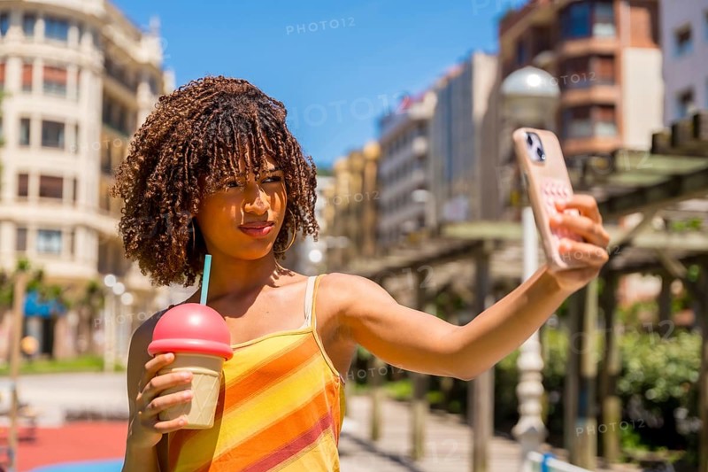 Young woman taking a selfie with her smartphone while holding a milkshake, enjoying a sunny day in the city during her summer vacation