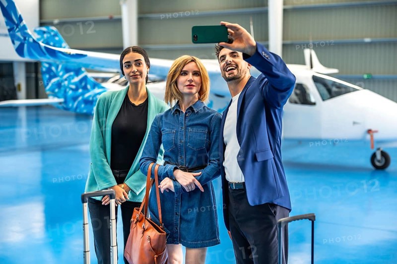 Three businesspeople are taking a selfie with a smartphone in a hangar with a private jet and luggage