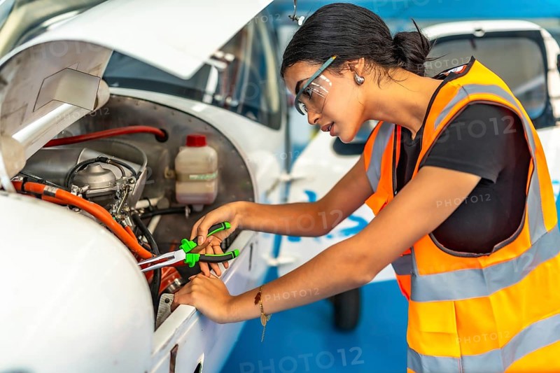 Young woman aircraft mechanic wearing safety glasses and high visibility vest using pliers while repairing airplane engine in hangar