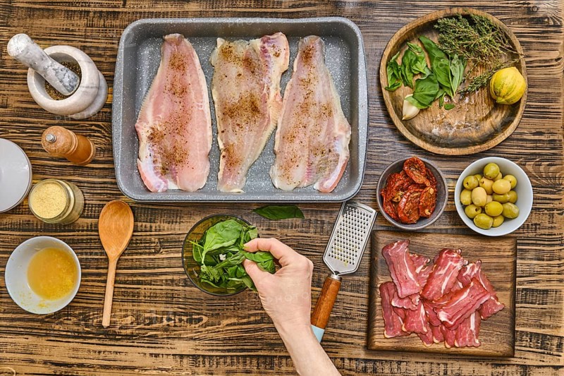 Fresh herbs, vegetables, and fish fillets are arranged on a wooden table. A hand adds spinach to a bowl while spices and garnishes await the next steps in crafting a delicious meal