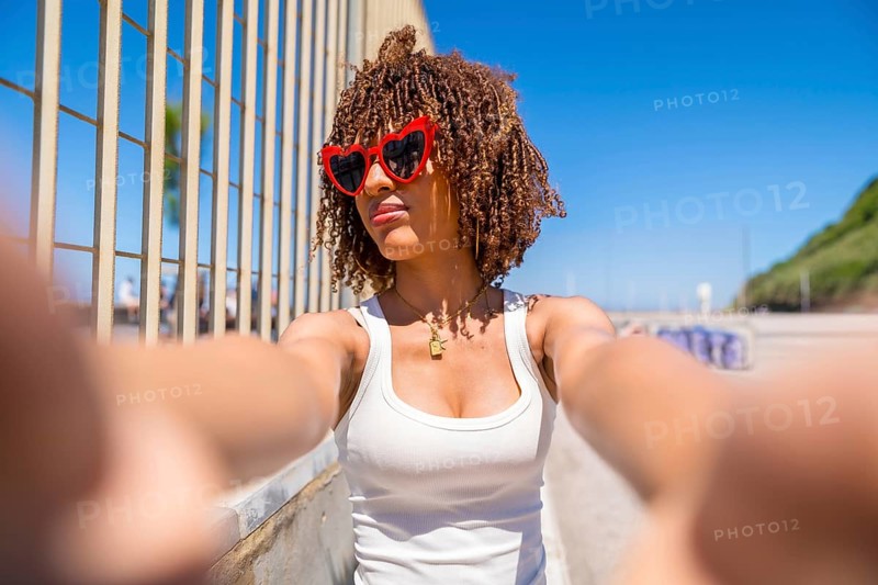 Portrait of a young woman with curly hair taking a selfie wearing heart shaped sunglasses by the beach