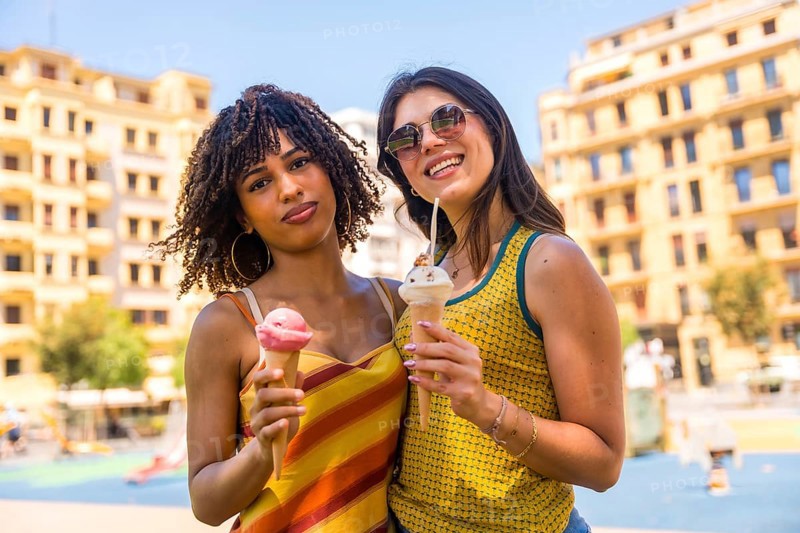 Two female friends enjoying ice cream cones together on a sunny summer day in a city park, sharing a moment of happiness and refreshment
