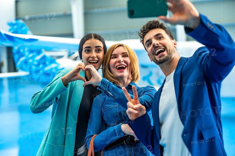 Three happy friends are taking a selfie in a hangar, making heart and victory signs, celebrating their private jet purchase