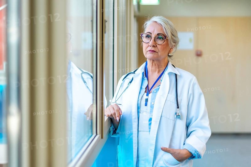 Thoughtful senior female doctor wearing lab coat and stethoscope looking through window in hospital corridor