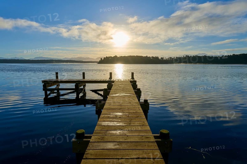 A jetty leads into a lake, the sun shines brightly and is reflected in the water, Lake Mahinapua, Ruatapu, South Island, New Zealand, Oceania