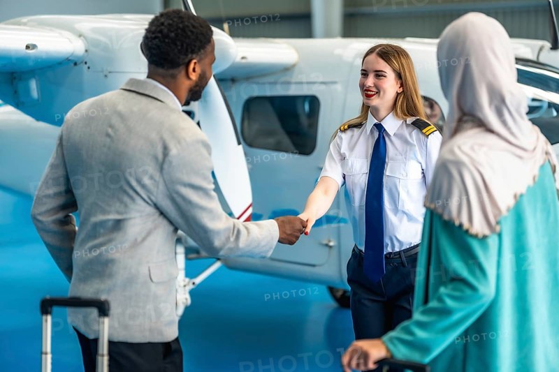 Female pilot shaking hands with businessman next to a private airplane inside an hangar, a businesswoman is waiting next to them