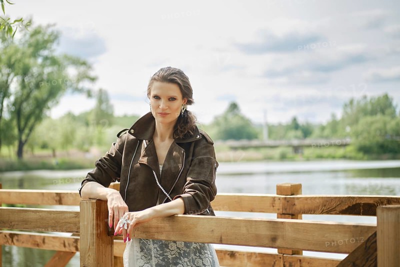 A woman stands confidently by a wooden railing in an elegant outfit, enjoying a peaceful afternoon by the lake with lush greenery and soft clouds in the background