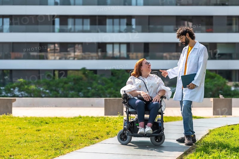 Doctor talking with smiling disabled woman in wheelchair, enjoying sunny day in urban park near hospital, promoting accessibility and healthcare