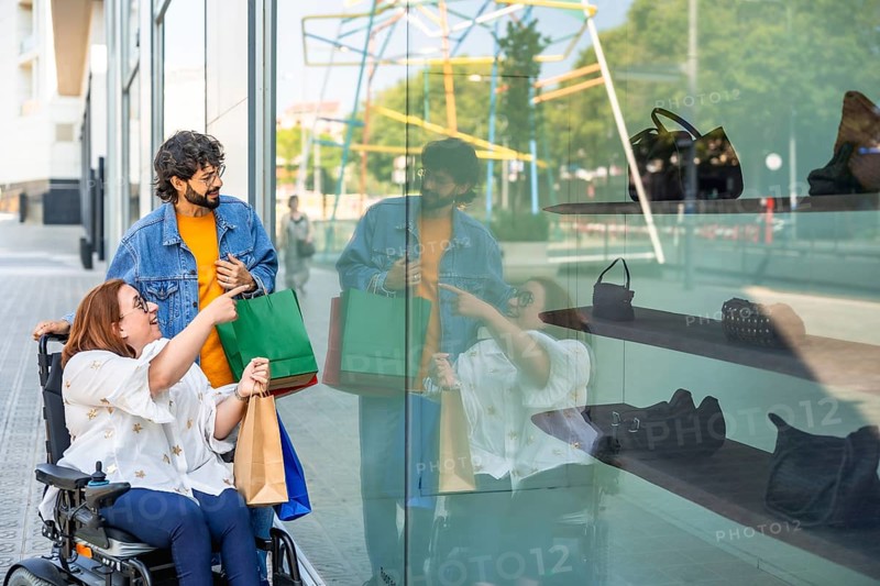 Happy friends doing shopping and looking at a shop window, a disabled woman on a wheelchair is pointing at a bag