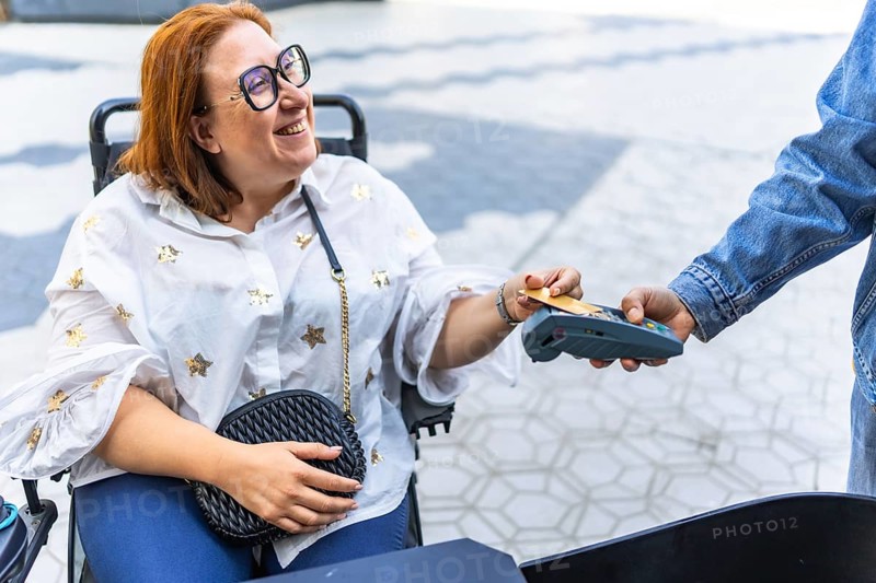 Happy woman using a credit card on a pos terminal while sitting in her wheelchair, enjoying a seamless outdoor shopping experience