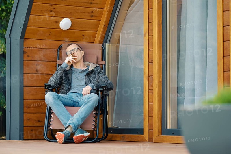 Middle-aged man sitting in lounge chair on the terrace of a modern tiny house with cihgar
