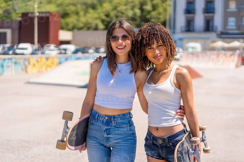 Two young women smiling and holding skateboards, enjoying their time together in a vibrant skatepark under the sunny summer sky