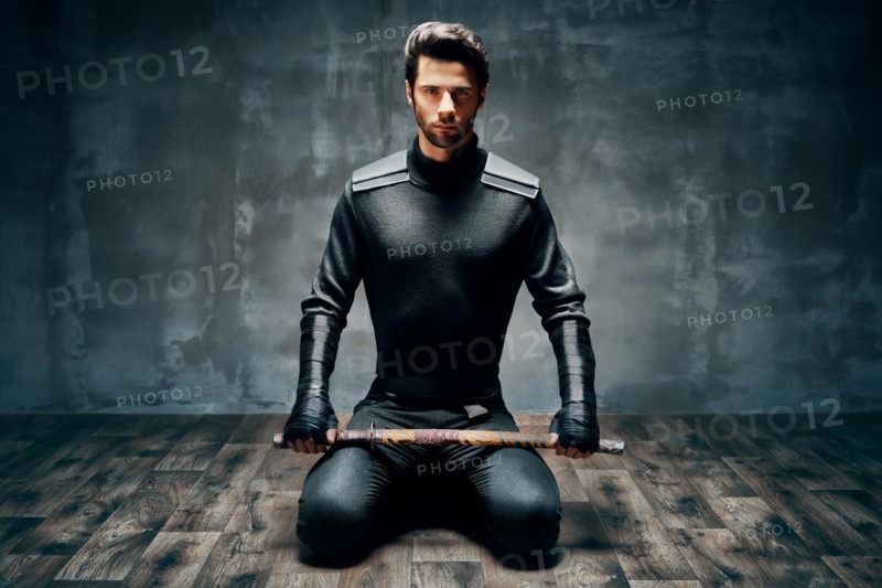 Warrior man sitting on floor posing with a sword over black background