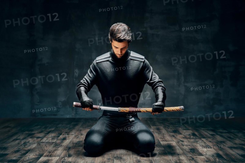 Warrior man sitting on floor posing with a sword over black background
