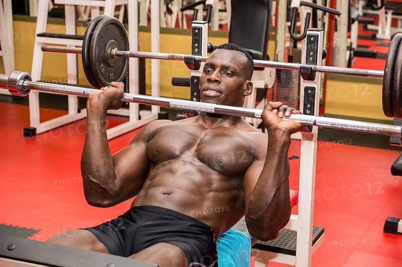 Hunky muscular black bodybuilder working out in gym, exercising pecs on bench with barbell