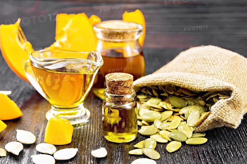 Pumpkin oil in vial, gravy boat and a jar, seeds in a bag and on table, slices of vegetable on black wooden board background