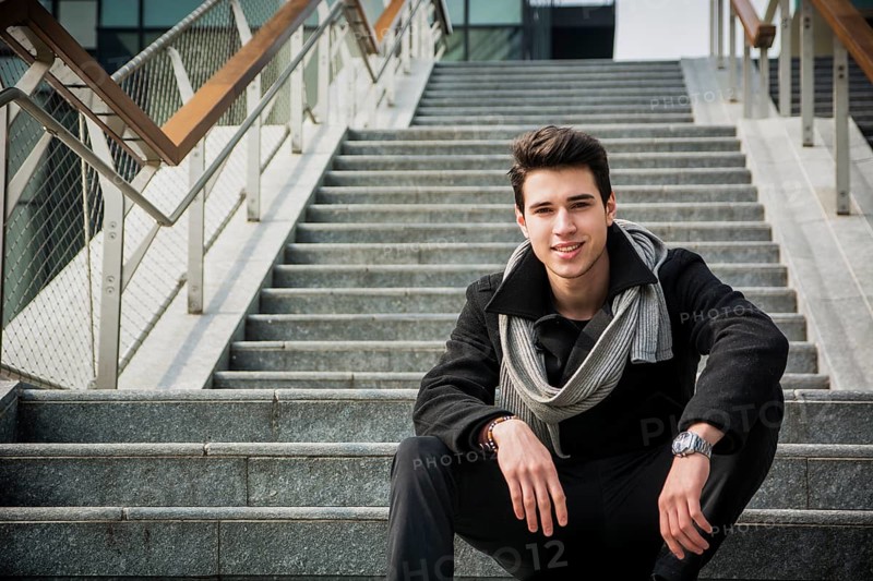 Trendy handsome young man in winter fashion sitting on a long staircase leading to an urban commercial building looking at the camera smiling