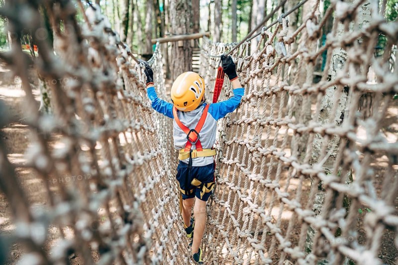 Boy walks across a mesh bridge, holding onto a cable. Back view.
