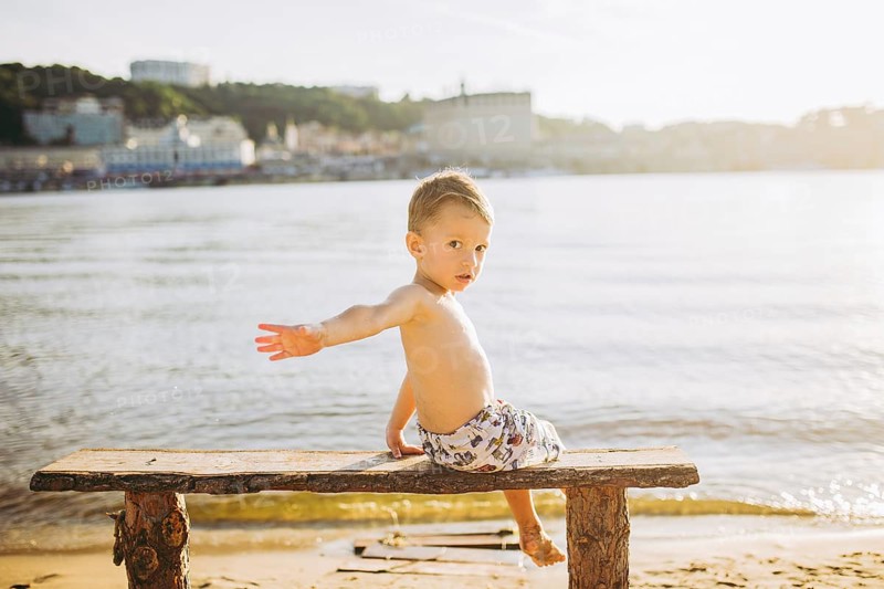 Boy child sitting on a wooden bench with his back on the beach near the water and showing his hand forward direction against the backdrop of the city of Kiev