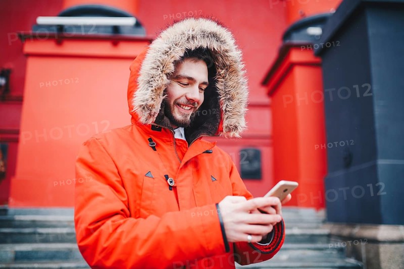 Handsome young male student with toothy smile and beard stands on red wall background, facade of educational institution in red winter jacket with hood with fur, Uses finger on screen of mobile phone