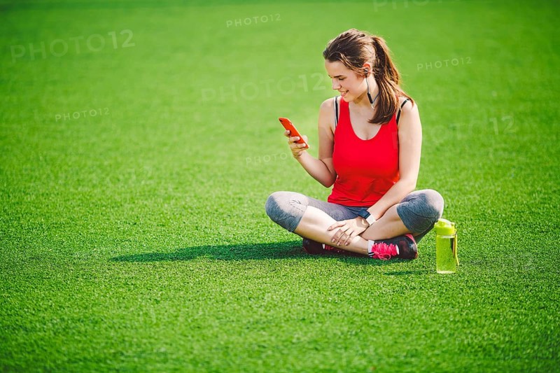 Theme sport health. beautiful young girl sitting resting on green grass lawn stadium using technodogies. In handphone in ear headphones listens music, summer sportswear smart watch heart rate monitor
