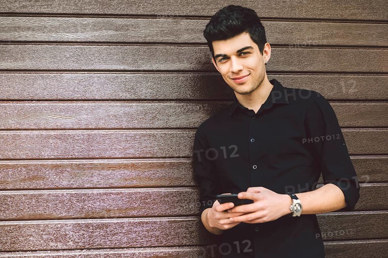 Portrait of young handsome Turkish male model. Mediterranean race brunette in black shirt is using a hand phone of a technology businessman on the street in sunny weather on a wooden wall background