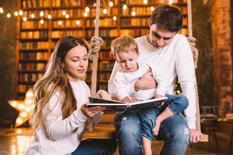 Family togetherness. Mom, dad and son reading story book together sitting on swing evening at home. Family and Parenthood Concept. Happy family reads childrens book while sitting home in loft interior