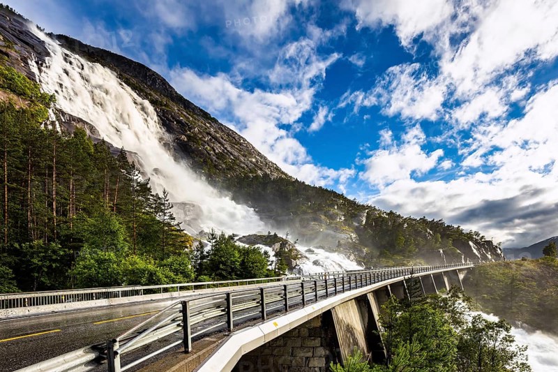 Huge powerful and roaring cascading Langfossen waterfall on the Vaule river. The bridge is fenced with railings. Passes through the highway. Summer cold and rainy day. Western Norway