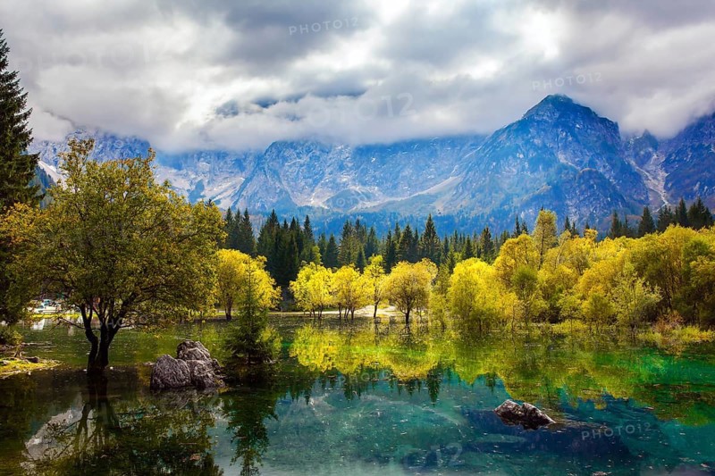 The Dolomites are covered with clouds. Lake Fuzine in Northern Italy. Magnificent colors of autumn. Orange and yellow trees are reflected in the green smooth water