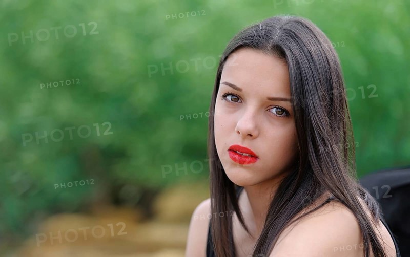 Young beautiful serious brunette girl with red lips and long straight hair over green nature background