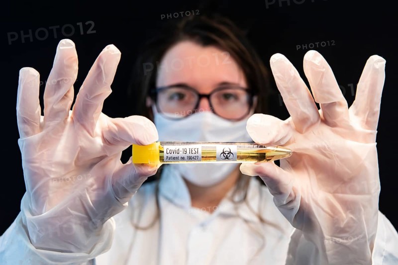 Selective focus shot of vial filled with urine sample and labeled Covid-19 test, held center frame by masked out of focus doctor over black background