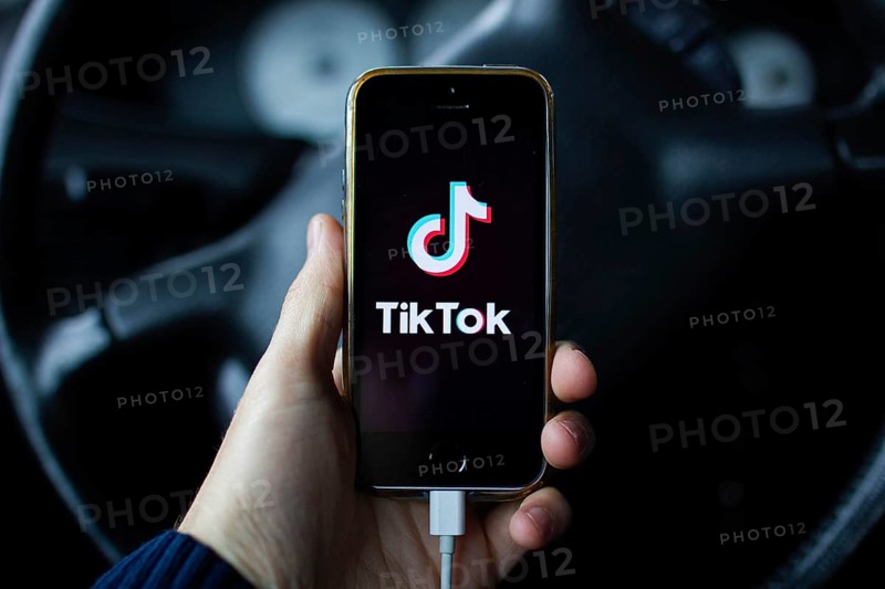 A hand holding a smartphone with TikTok app in front of a steering wheel inside a car