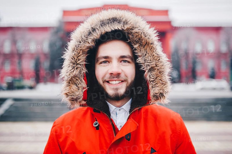 Portrait, close-up of young stylishly man smiling with a beard dressed in red winter jacket with a hood and fur on his head stands against the background of a snow-covered city. Winter and frost theme