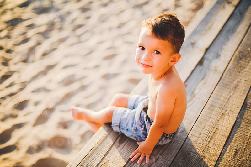 Little Caucasian boy child sitting on wooden pier sandy beach, summer time, sea holiday near water. The theme is the flow of time, a short life, the meaning and purpose of existence. Meaning of life
