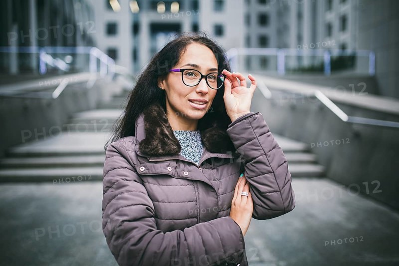 Portrait beautiful young business woman brunette in jacket and jumper stands on background office building, business centre in glasses for view. Hand holding onto shackle of glasses and looking away